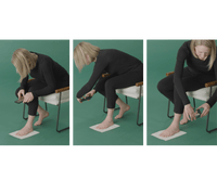 A seated woman using her smartphone to scan each foot on a sheet of printer paper, the guided steps for getting IAMBIC precision-fit shoes.