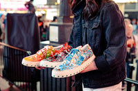 A person holding three hand-painted Art Gods x IAMBIC sneakers in Times Square.