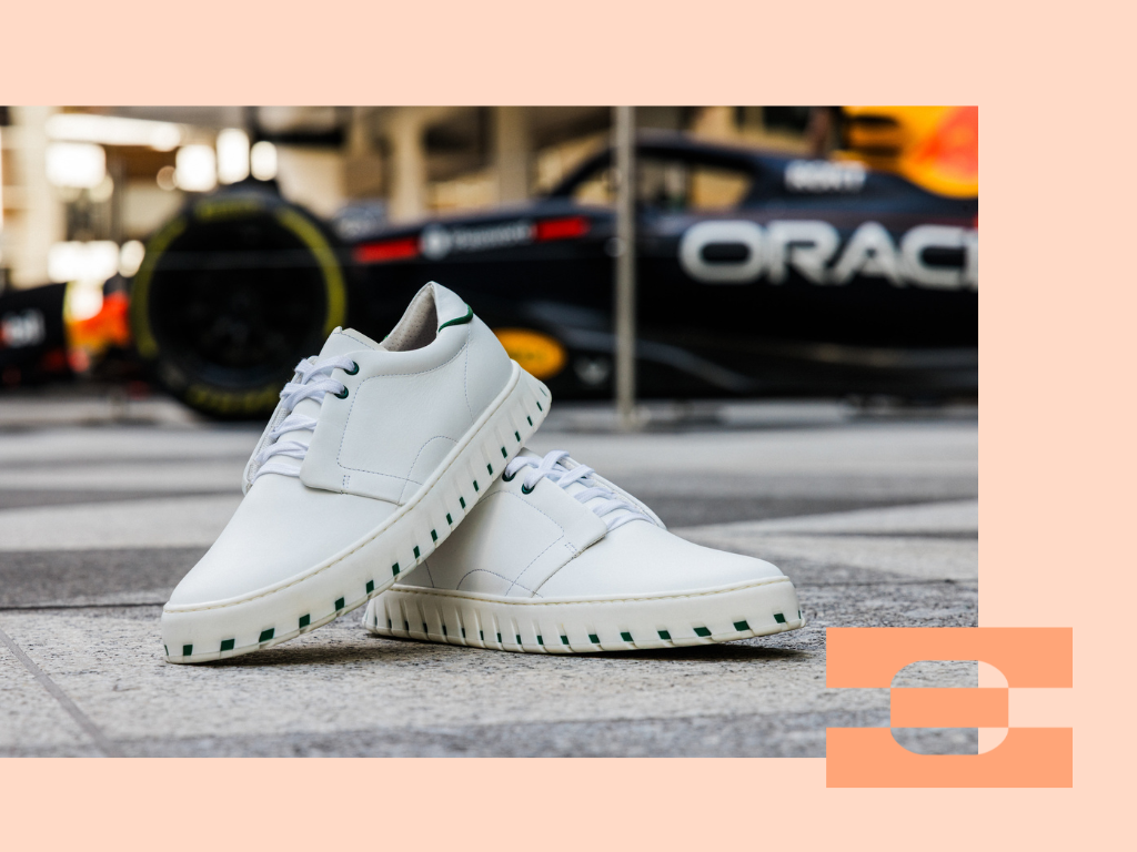 Two white IAMBIC MODEL T custom-fit shoes on stone pavement with a blurred race car in the background.